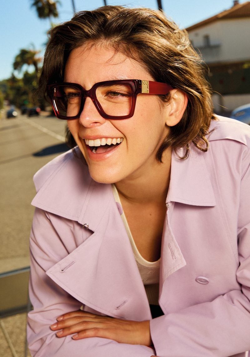 Modella sorridente con occhiali da vista bordeaux MAX&Co. in trench rosa